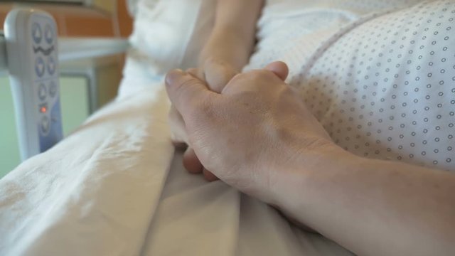 Support person in difficult times. Husband holds the hand of his wife, who lies in the hospital cot. Patient after surgery regains consciousness. Emotional moment in close-up