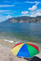 bunter Sonnenschirm am Gardasee Strand, Segelboot auf dem See und Berge im Hintergrund