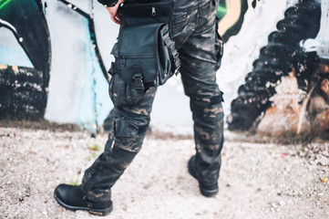 An urban man in a camouflage military pants stands next to the wall. Background from the urban part of the city