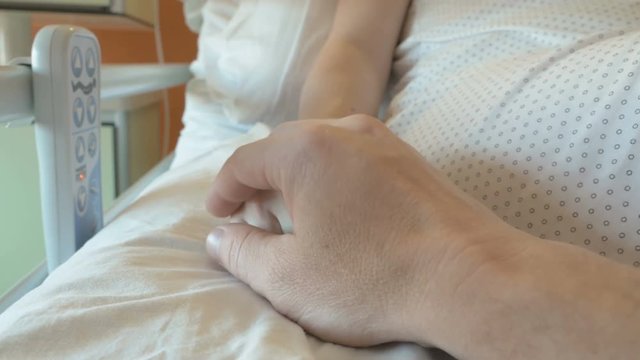Woman after surgery feels a lot of pain. Husband holds the hand of his wife, who lies in the medical bed. Support for a woman after giving birth in hospital ward. Emotional moment in close-up