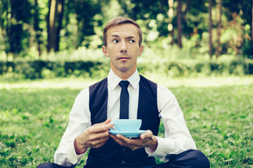 Young attractive man in a vest and tie  resting on the nature and drinking coffee. Holds a cup of tea.