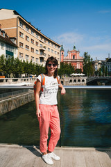 Fototapeta na wymiar Young smiling woman tourist standing on a bridge in the old centre of Ljubljana, Slovenia, wearing white shirt, white sneakers and coral trousers. With an orange backpack. Travel concept.