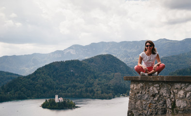 Young smiling woman tourist sitting on a Bled Castle wall with the lake Bled and the Bled island on the background. Surrounded by mountains and forests in Slovenia. Travel concept.