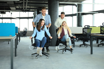 Fototapeta premium Young cheerful business people in smart casual wear having fun while racing on office chairs and smiling.
