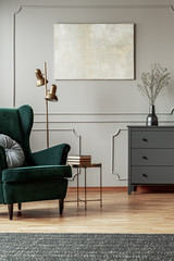 Emerald green wing back chair with pillow in grey living room interior with wooden commode