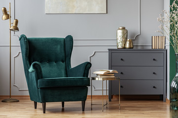 Books on stylish golden small table next to emerald green velvet wing back chair in grey living...