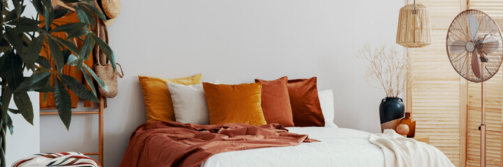 Real photo of a cozy bedroom interior with colorful pillows, fan and close-up of a plant