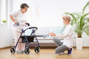 Senior woman with walker trying to walk again and helpful physiotherapist supporting her - obrazy, fototapety, plakaty