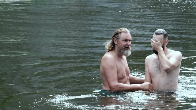 A grown man with a beard, like a priest, dips a young bearded guy into the water. The concept of baptism in the river or entertainment on vacation. People and nature, religion and lifestyle.