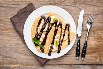 French crepe with banana and chocolate sauce on white plate, wooden background. Table top view