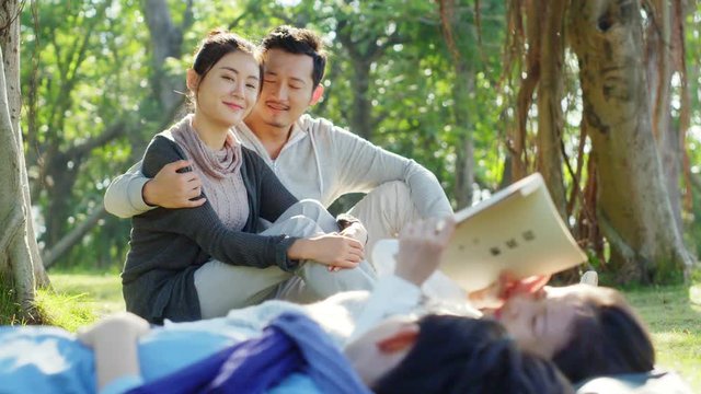 asian parents watching two children lying on grass reading a book together in park