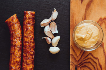 Two sausages and garlic next to wooden board with a bowl of mustard on it – Pork and cloves next to seasoning on black ardesia stone