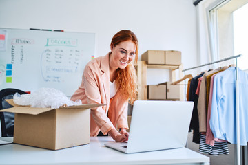 Small business concept. Woman looking at laptop while packing sold items in office