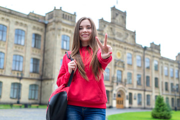 Low angle photo of beautiful pretty showing v-sign to you in camera holding modern schoolbag. Old building is on the background