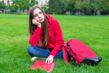 Photo of depressed passive sad teen girl sitting on green grass lawn is not willing to do home task...