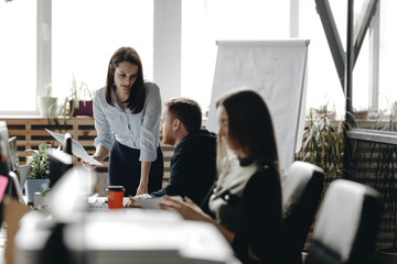 Young brunette girl dressed in office style clothes is sitting at the desk with the colleagues and keeping the notebook in her hands in a light modern office