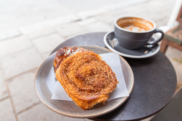 Warm cinnamon bun with icing sugar with a cup of coffee lying on the tablein a street cafe.