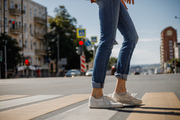 Young girl dressed in jeans and sneakers is crossing the road with a backpack in a city street on a summer sunny day