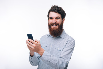 Photo of handsome cheerful young man with beard holding smartphone and looking at the camera over...
