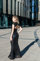 Fototapeta premium A beautiful blonde girl stands with her back in a black evening long dress against the backdrop of a modern building.