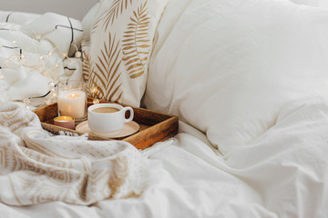 Fototapeta na wymiar Wooden tray of coffee and candles on bed. White bedding sheets with striped blanket and pillow. Breakfast in bed. Hygge concept.