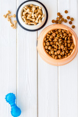 Dry pet food in bowls and dog toys, care tools on white wooden background top view space for text