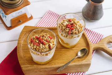 Granola with mango sauce and goji berries. On a wooden board, an iron spoon, a coffee pot, a white wooden table and a napkin.