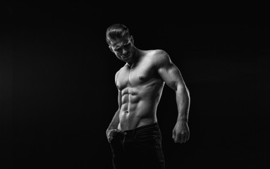 Naklejka na ściany i meble Black and white. Muscular model sports young man on dark background. Fashion portrait of strong brutal guy with a modern trendy hairstyle. Sexy torso. Male flexing his muscles.