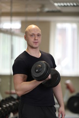 Fototapeta na wymiar european caucasian athletic man bodybuilder in black sportwear holding dumbell and showing his muscular arms. man doing exercise for biceps. Background is gym