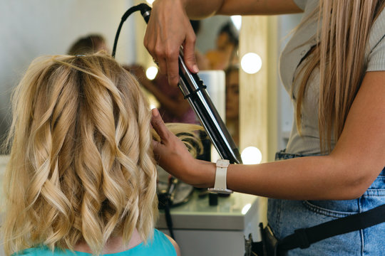 Hairtician twirls curls with flat iron hair. Back view. Hairdressing services. Creation of evening hairstyles fashionable stylish women's hairstyles. Hair styling process. Courses in hairdressing. Bea