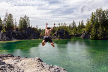 Athletic and Adventurous Man is Cliff Jumping into a Green Colored Glacier Lake during a hot and sunny summer day. Taken in British Columbia, Canada.