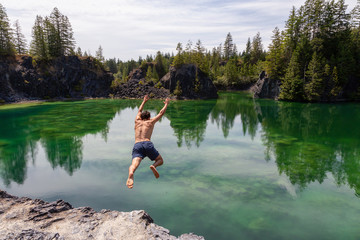 Athletic and Adventurous Man is Cliff Jumping into a Green Colored Glacier Lake during a hot and sunny summer day. Taken in British Columbia, Canada.