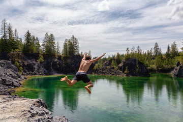 Fototapeta na wymiar Athletic and Adventurous Man is Cliff Jumping into a Green Colored Glacier Lake during a hot and sunny summer day. Taken in British Columbia, Canada.