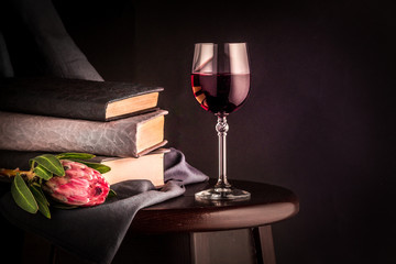 A glass of red wine, a pink protea flower and a book against a dark background. Still life with flowers. Free space for your text.