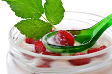 Pink cream or milk fruit yogurt with natural ripe red raspberry berry and green raspberry leaves in open glass transparent jar and curved slim graceful spoon on white background close up. Isolated