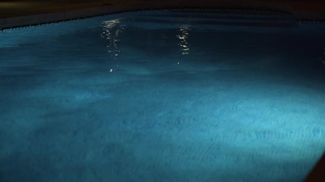 A swimming pool at night reflecting the pool side lights gently on the surface while illuminated by the inner pool light.