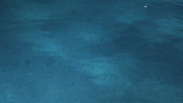 A swimming pool at night illuminated below the surface by the pool light with slight reflections of the poolside lights on the water's surface.