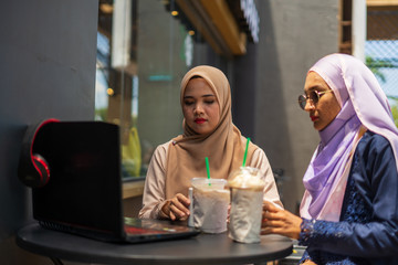 two happy young Asian Muslim women in coffeehouse or coffee shop, discussing each other, leisure time with friends with laptop.