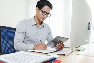 Young serious entrepreneur reading report on tablet computer and taking notes in planner