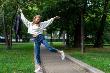 Fototapeta premium beautiful young European girl walks in city green park. Caucasian girl posing standing on one leg. Lifestyle, pullover, jeans, sneakers. interesting pose full length in fashionable look.