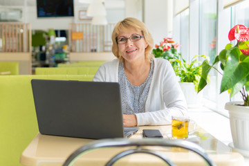A middle-aged woman with a laptop works in a cafe in the office, she is a freelancer. A woman with glasses sits at a table with a Cup of herbal tea, looks on camera and smiles.