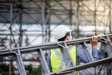 Asian maintenance worker man with safety helmet and green vest carrying aluminium step ladder at construction site. Civil engineering, Architecture builder and building service concepts © zephyr_p