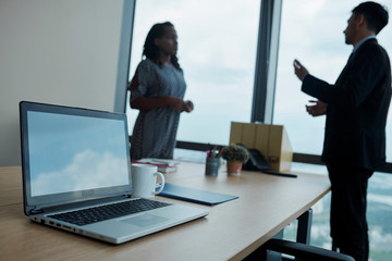 Opened laptop and mug of coffee on office table, talking business people in background