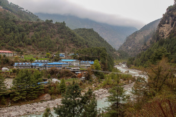 Phakding village stands on the riverbank of Dudh Koshi snow river (Dudh Kosi river or Milk-Koshi River) in Nepal. Suspension bridge connects two riverbanks. Nature, Countryside and Travel concept. - obrazy, fototapety, plakaty