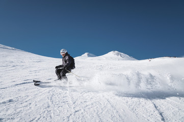 Naklejka na ściany i meble A bearded mature aged male skier in a black ski suit descends along the snowy slope of a ski resort amid two peaks of Mount Elbrus. The concept of sports in adulthood