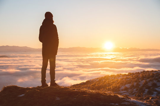 View From the back. A lonely standing man high in the mountains looks at the setting sun and the sunset horizon with a valley filled with clouds. The concept of tourism travel and male loneliness