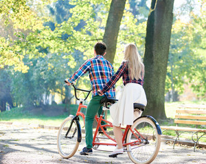 Obraz premium Back view of young active traveler couple, handsome man and attractive blond woman cycling together tandem double bike along crackled path in lit by bright sun beautiful park under tall trees.
