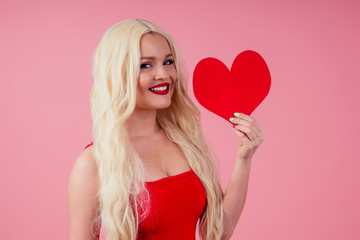 Closeup portrait of beautiful blond girl holding in hands paper heart isolated on pink background, Valentine day celebration