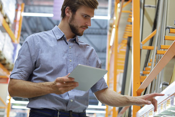 Young man shopping or working in a hardware warehouse standing checking supplies on his tablet.