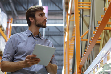 Young man shopping or working in a hardware warehouse standing checking supplies on his tablet. © ty